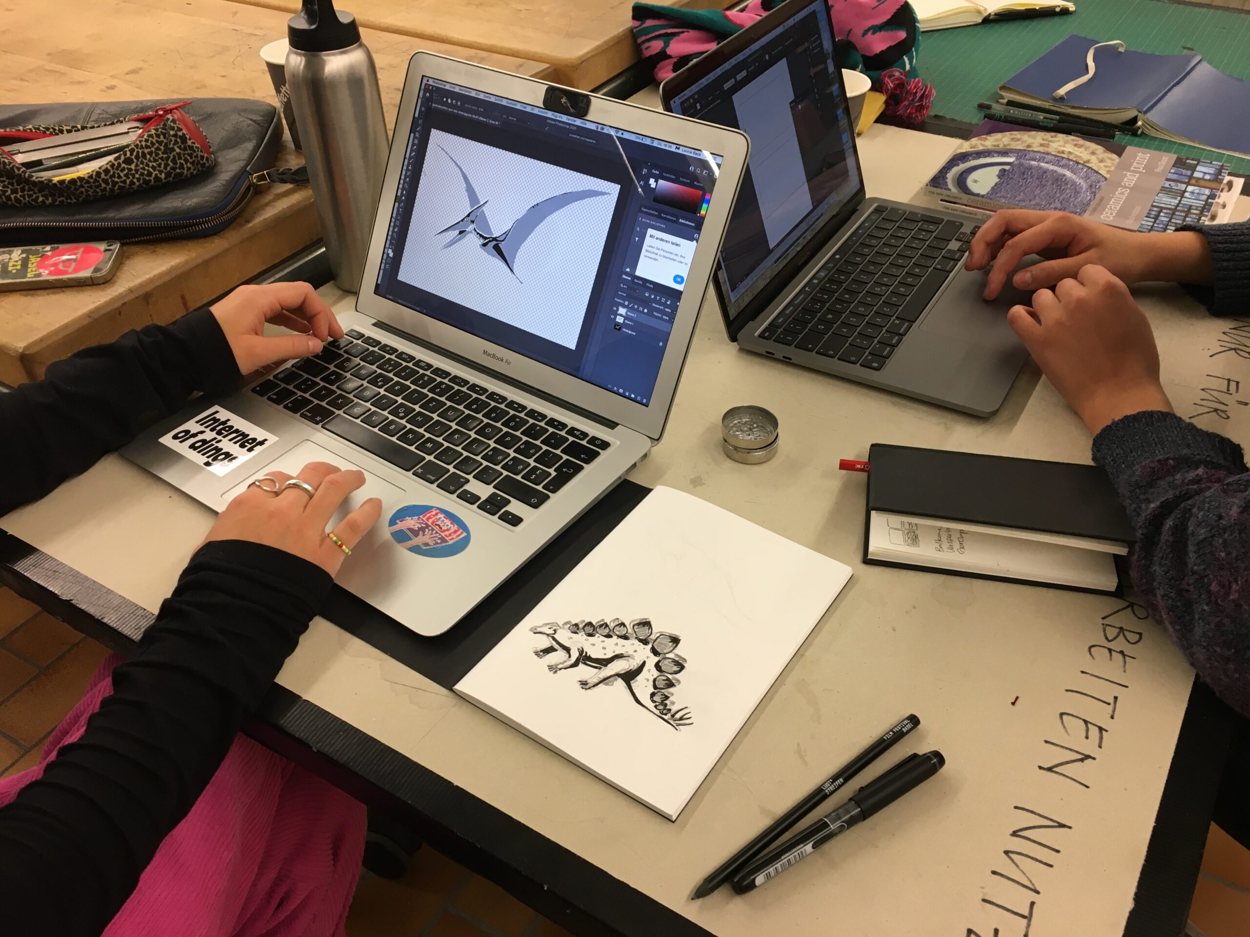 Két nyitott laptop áll egy asztalon, és egy-egy személy dolgozik rajta. Kezük a billentyűzeten pihen. A képernyőn egy repülő őshüllő (pteroszaurusz) grafikája látható. A képernyőtől jobbra egy vázlatfüzet hever, amelyben egy dinoszaurusz tussal készült rajza látszik.
