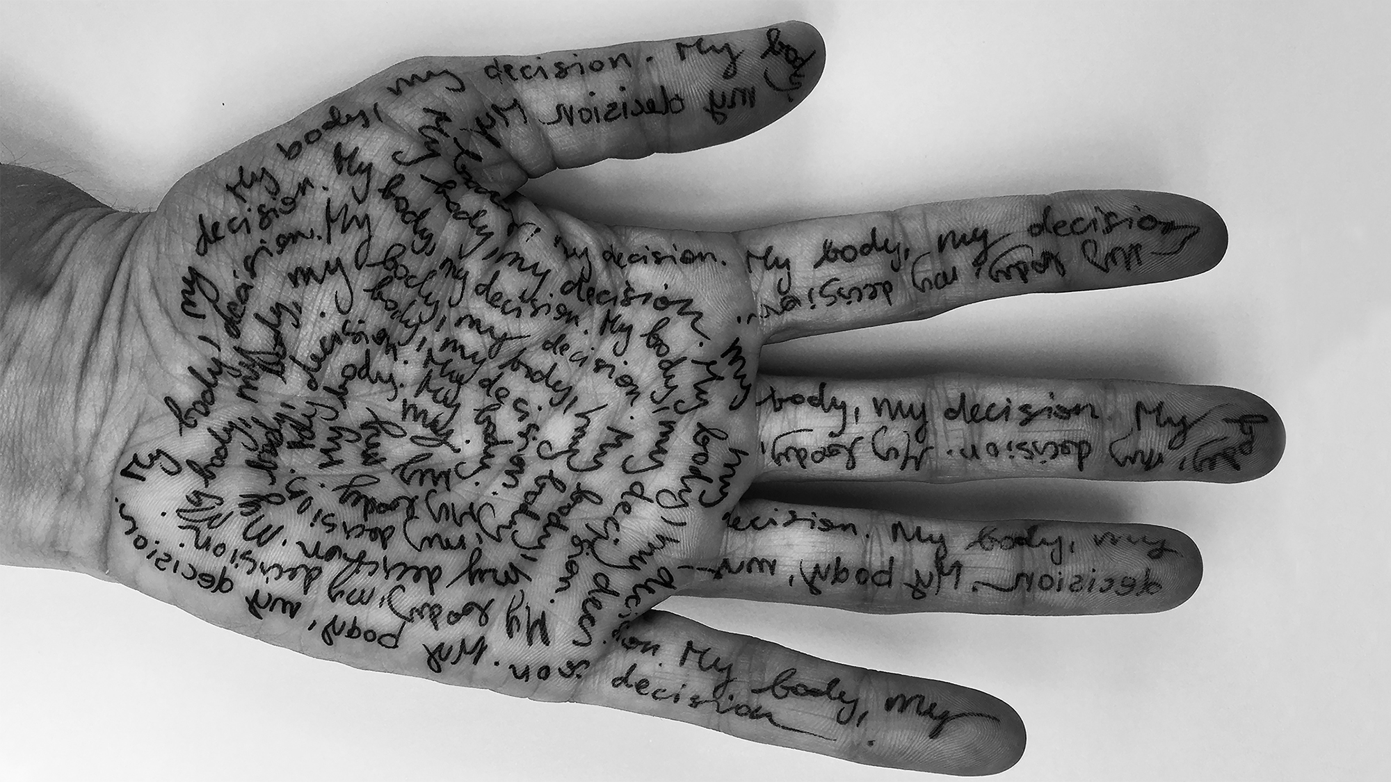 Eine Schwarzweiß-Fotografie zeigt eine mit kleinen Buchstaben beschriebene Handfläche, auf der wiederholt der Satz „My body, my decision“ zu lesen ist.