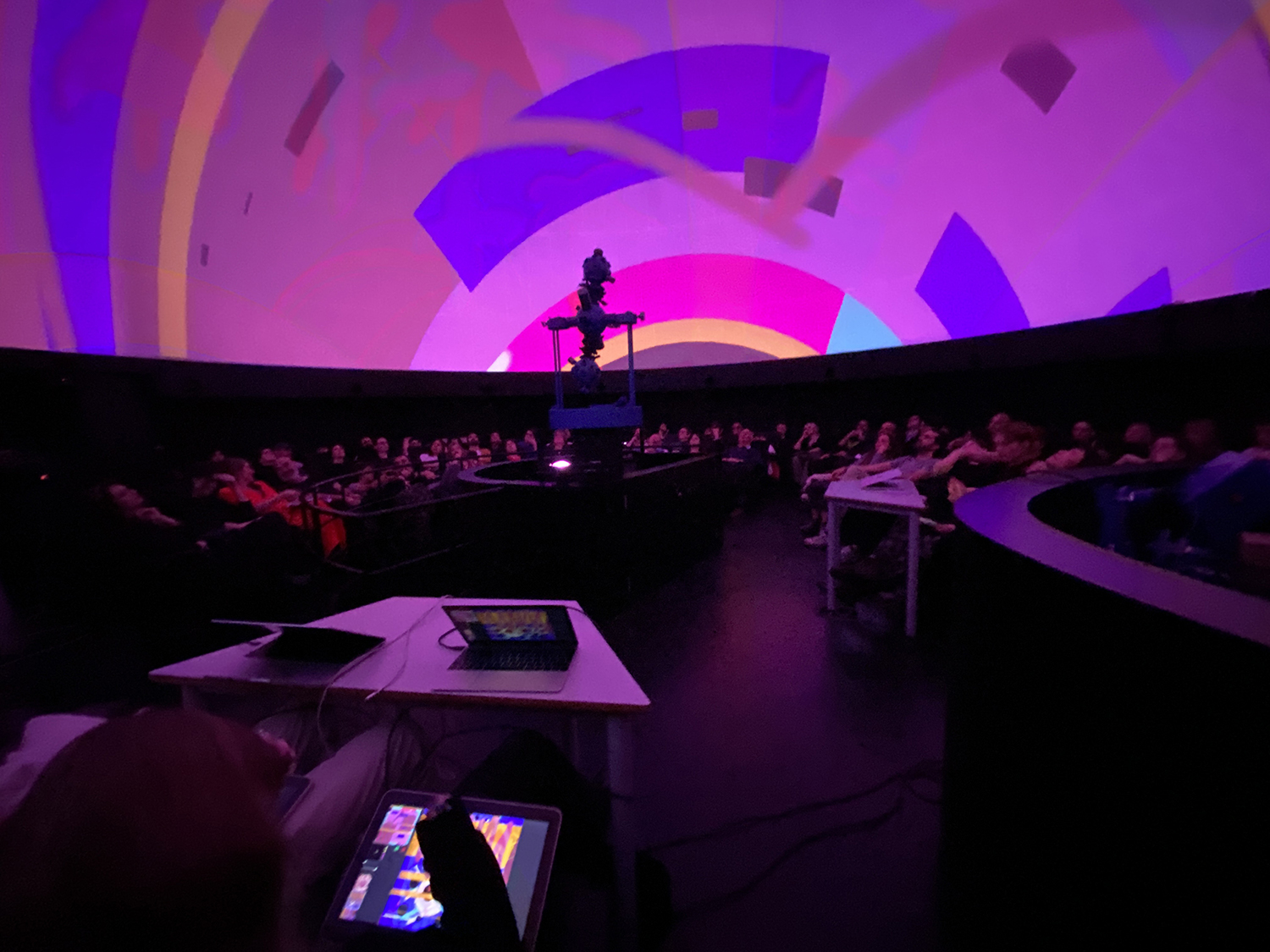 A dome-shaped auditorium filled with an audience sitting in dim lighting. The ceiling displays vivid, abstract projections in pink, purple, and yellow hues, creating a dynamic visual experience. A central device resembling a projector stands in the middle, surrounded by tables with laptops and tablets used for controlling the visuals.