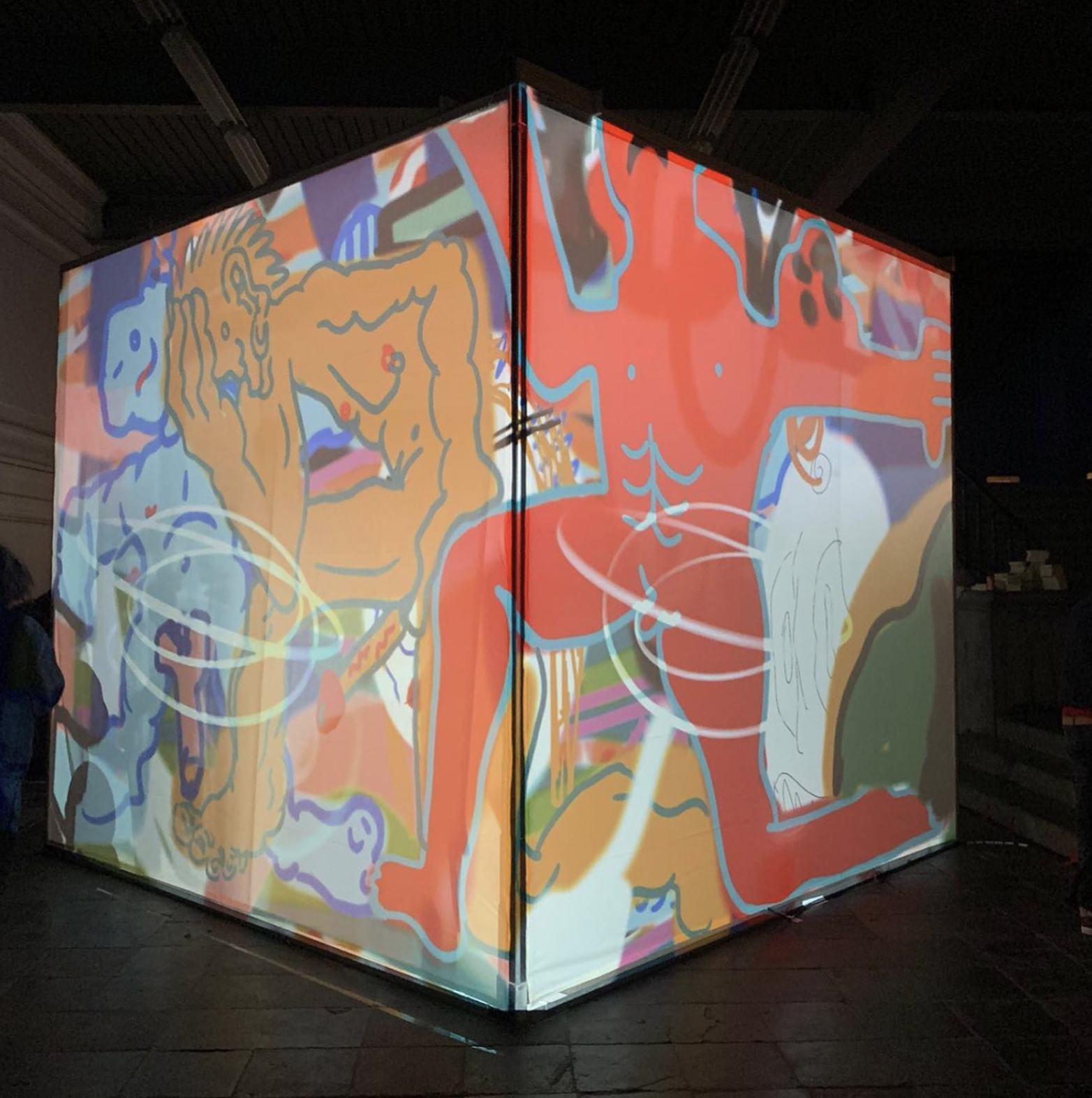 A large cube-shaped structure with projected illustrations on its surfaces. The projections depict abstract and colorful figures, including a red humanoid figure with exaggerated muscles and a seated orange figure with spiky hair. The background is filled with intricate and vibrant designs, creating a bold and immersive art installation.