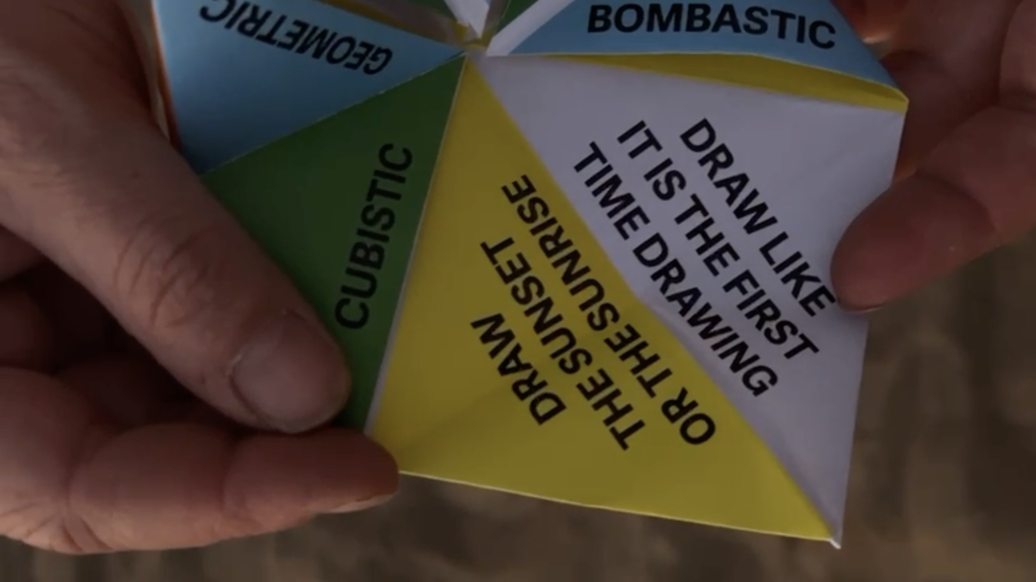  A close-up of a hand holding a folded paper fortune teller with colorful triangular sections. The visible sections have words such as 