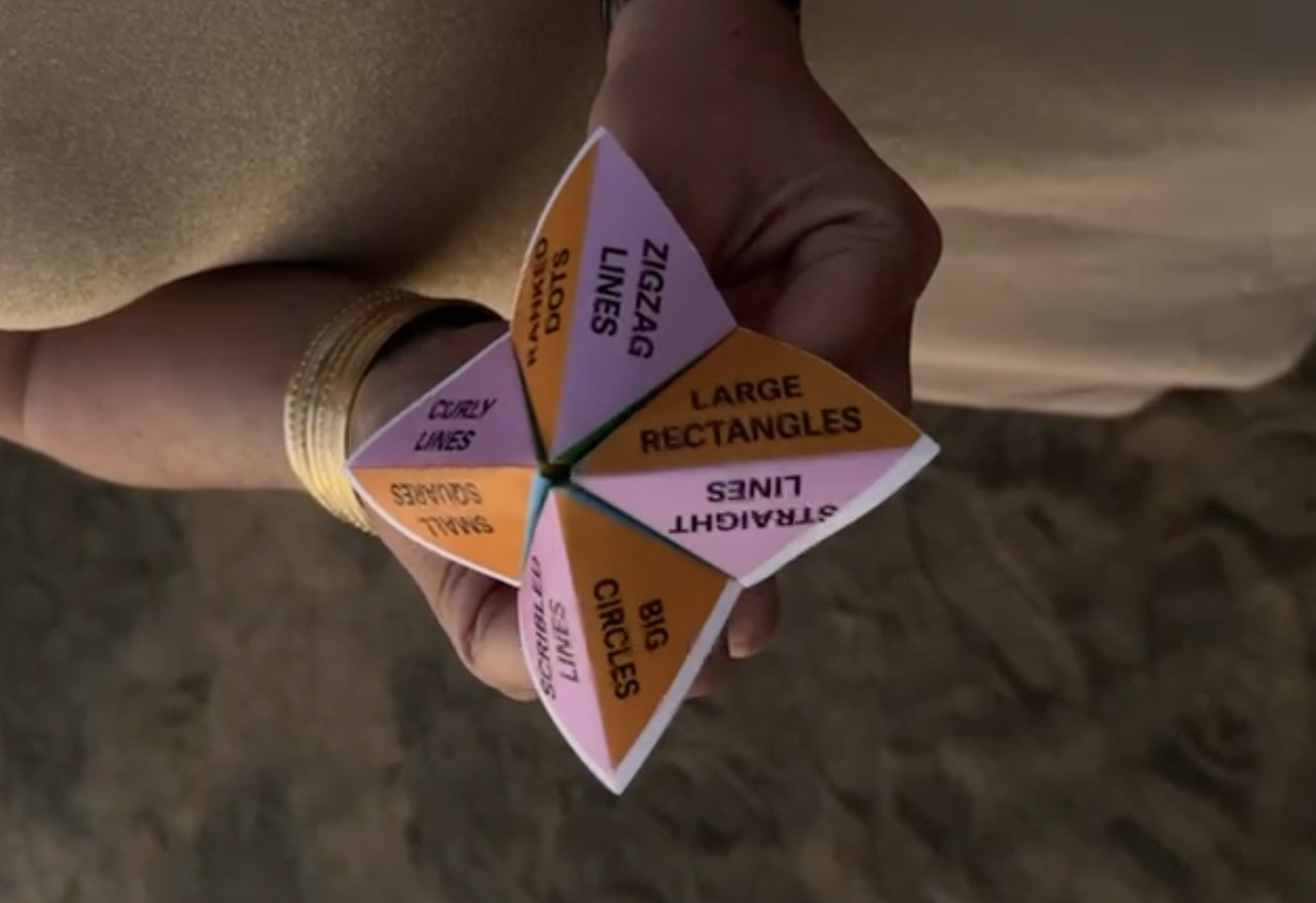  A close-up of another folded paper fortune teller held in a hand. The visible sections are labeled with drawing prompts, including 