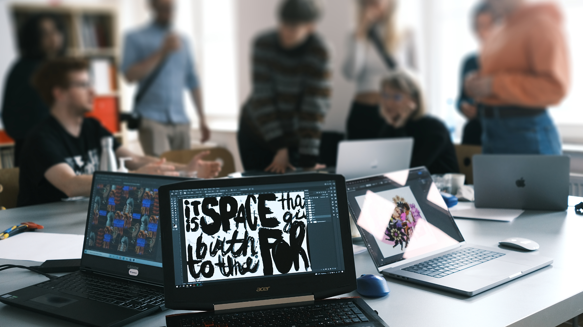 Een gezamenlijke werkruimte met een groep mensen verzameld rond een tafel. Op de voorgrond tonen verschillende laptops creatieve werken: een ervan toont de tekst 