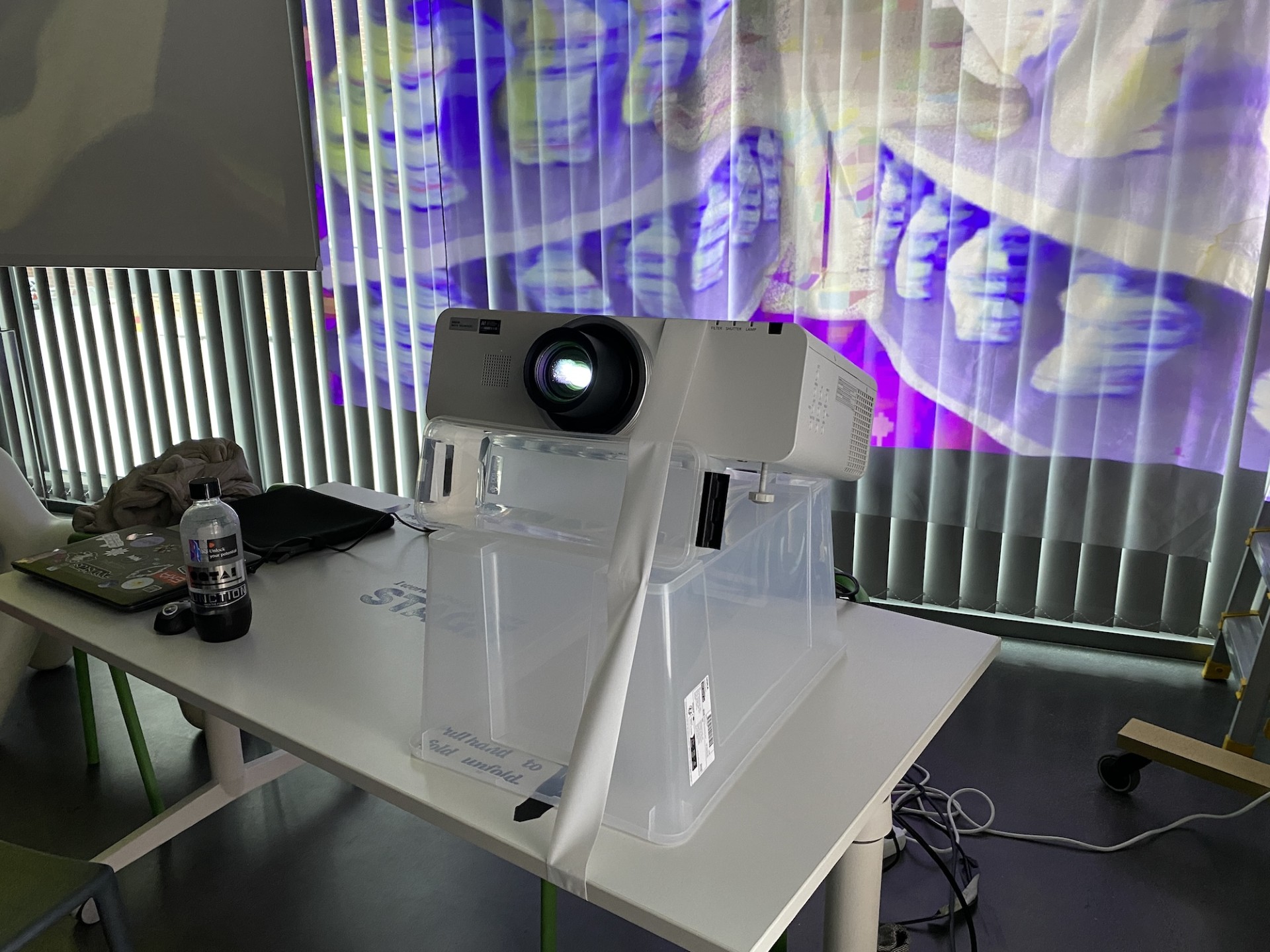 Een klaslokaal- of kantoorsetup met een projector die op een transparante plastic doos op een tafel staat. De projector is ingeschakeld en projecteert kleurrijke en abstracte paarse en gele visuals op verticale lamellen in de achtergrond. Op de tafel staan een laptop met stickers, een fles frisdrank en verschillende kabels en andere items, wat een experimentele of creatieve werkplek suggereert.