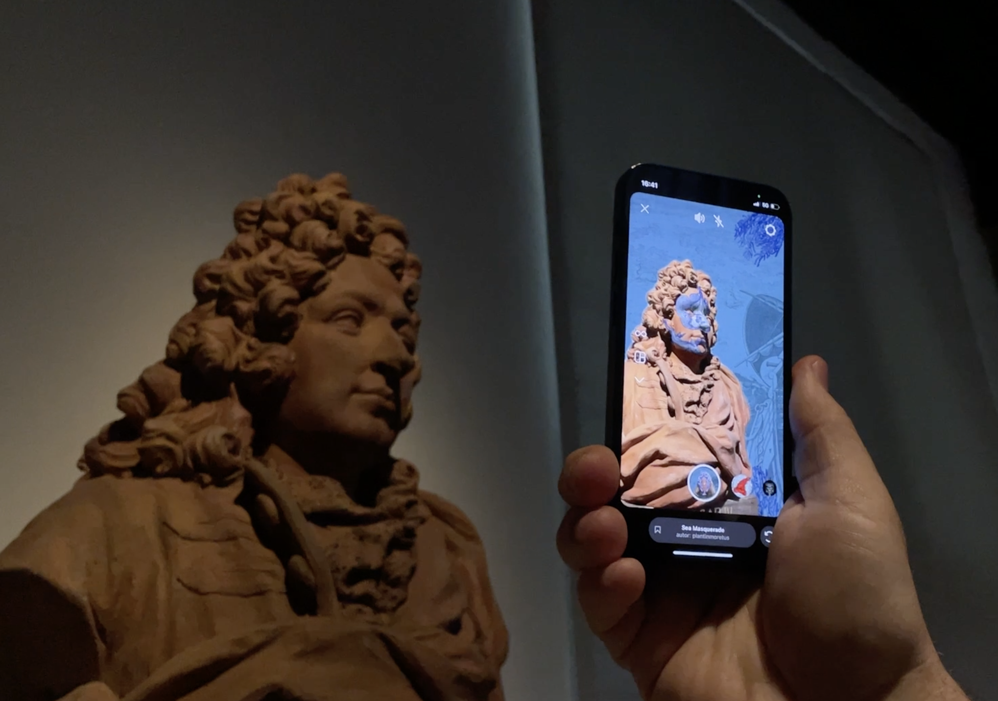 Een foto van een klassieke buste van een man, gedetailleerd met gekruld haar en een formele outfit, verlicht in een schemerige omgeving. Op de voorgrond houdt een hand een smartphone vast die een augmented reality-weergave van dezelfde buste toont. Op het scherm is de buste te zien met een digitale overlay en interactieve elementen.