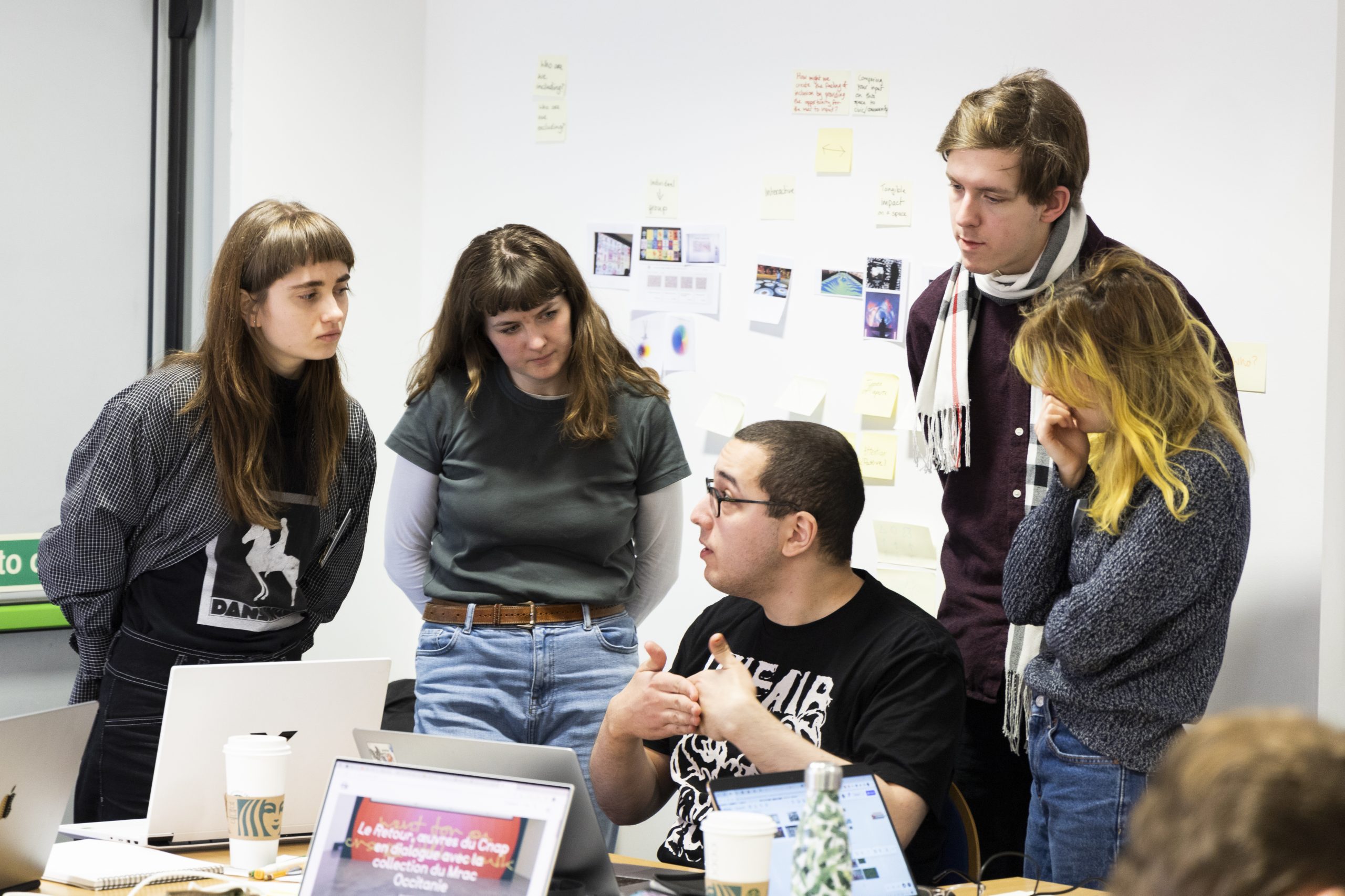 Een kleine groep mensen die in een gerichte discussie rondom een tafel zit, met laptops en aantekeningen. Een persoon aan tafel spreekt en gebaart, terwijl de anderen aandachtig luisteren. Op de achtergrond hangen plakkertjes en geprinte materialen aan de muur, wat wijst op een creatieve of planningssessie.