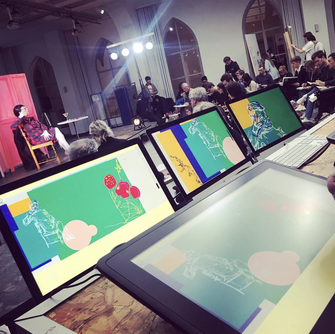 A digital art workshop with multiple monitors displaying colorful sketches of a person seated with abstract objects like mushrooms and geometric shapes. In the background, participants observe a live drawing session, where a model poses under studio lights.