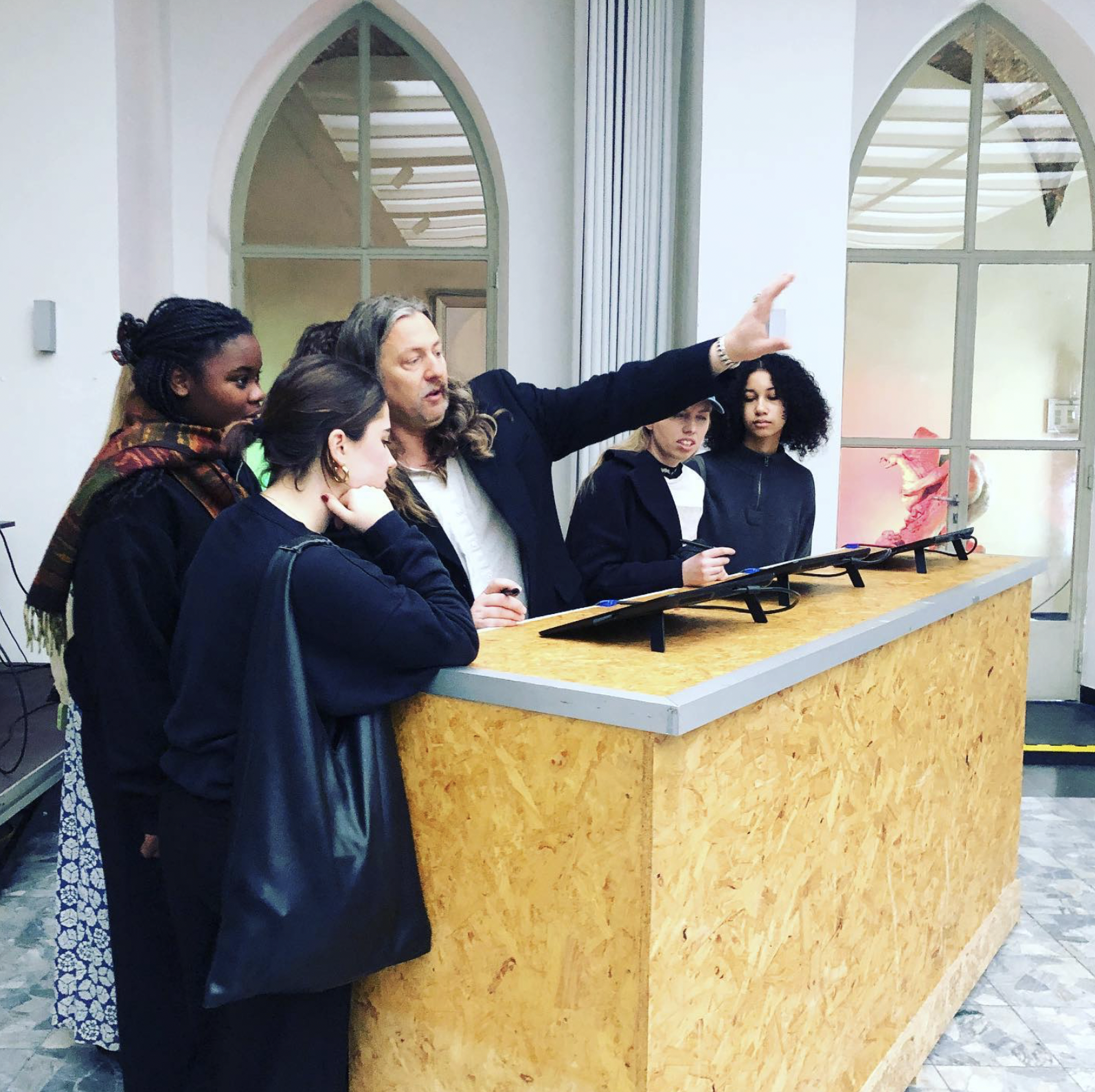 A group of people gathered around a wooden counter with digital screens displayed at an angle. A man with long hair gestures toward the screens, explaining something to the attentive group. The room has high arched windows, giving it a bright and spacious feel.