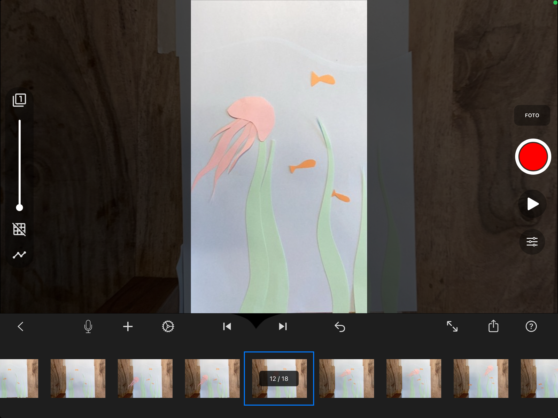 Auf dem Bild ist die Oberfläche der Stop-Motion-Studio App zu sehen. Im unteren Abschnitt befinden sich die einzelnen Fotografien, welche vom linken bis zum rechten Rand angeordnet sind, die dann abgespielt werden können. Auch die Bearbeitungstools, wie Beispielsweise die Frames per Second oder weitere Buttons für das Downloaden etc. findet man unten in der Leiste genau über den Fotografien. Rechts am Rand findet sich der Play Button und der Knopf, mit dem Fotografiert wird. Beispielhaft ist als zu fotografierendes Element ein blauer Hintergrund auf dem eine Wasserlandschaft mit Fischen, Algen und einer Qualle zu sehen ist.